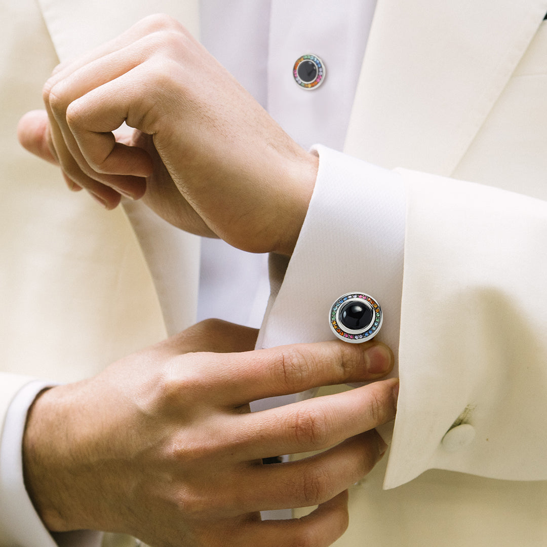 Man in ivory tuxedo adjusting black onyx and rainbow crystal cufflink on white French cuff with matching tuxedo stud visible.