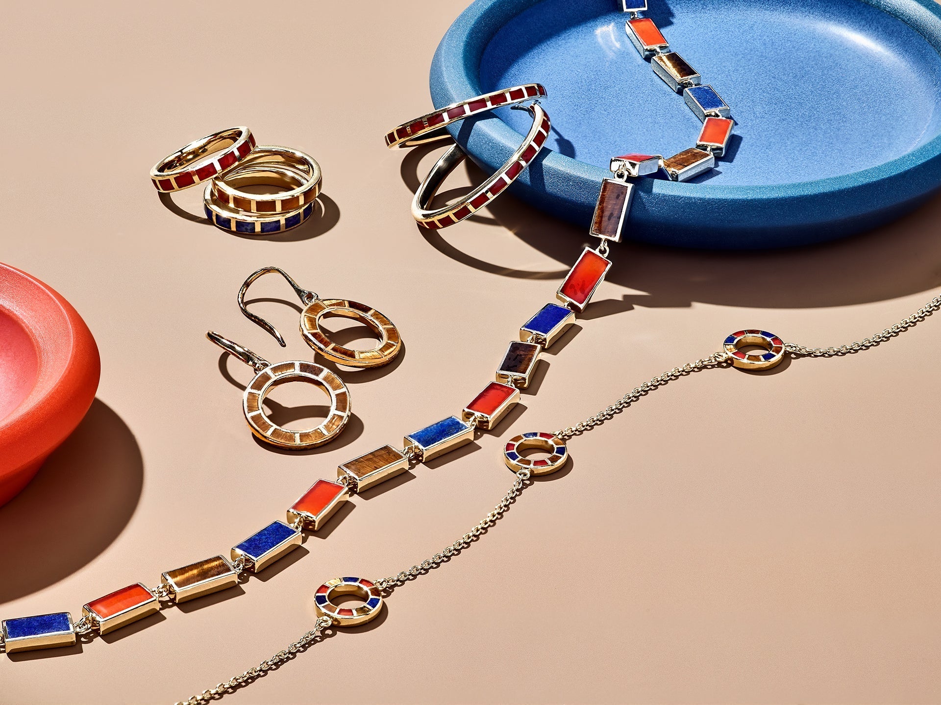Bold gold-plated mixed gemstone jewelry collection featuring lapis lazuli, tiger's eye, and carnelian inlay across stackable rings, open-circle earrings, a cuff bracelet, and a long station necklace.