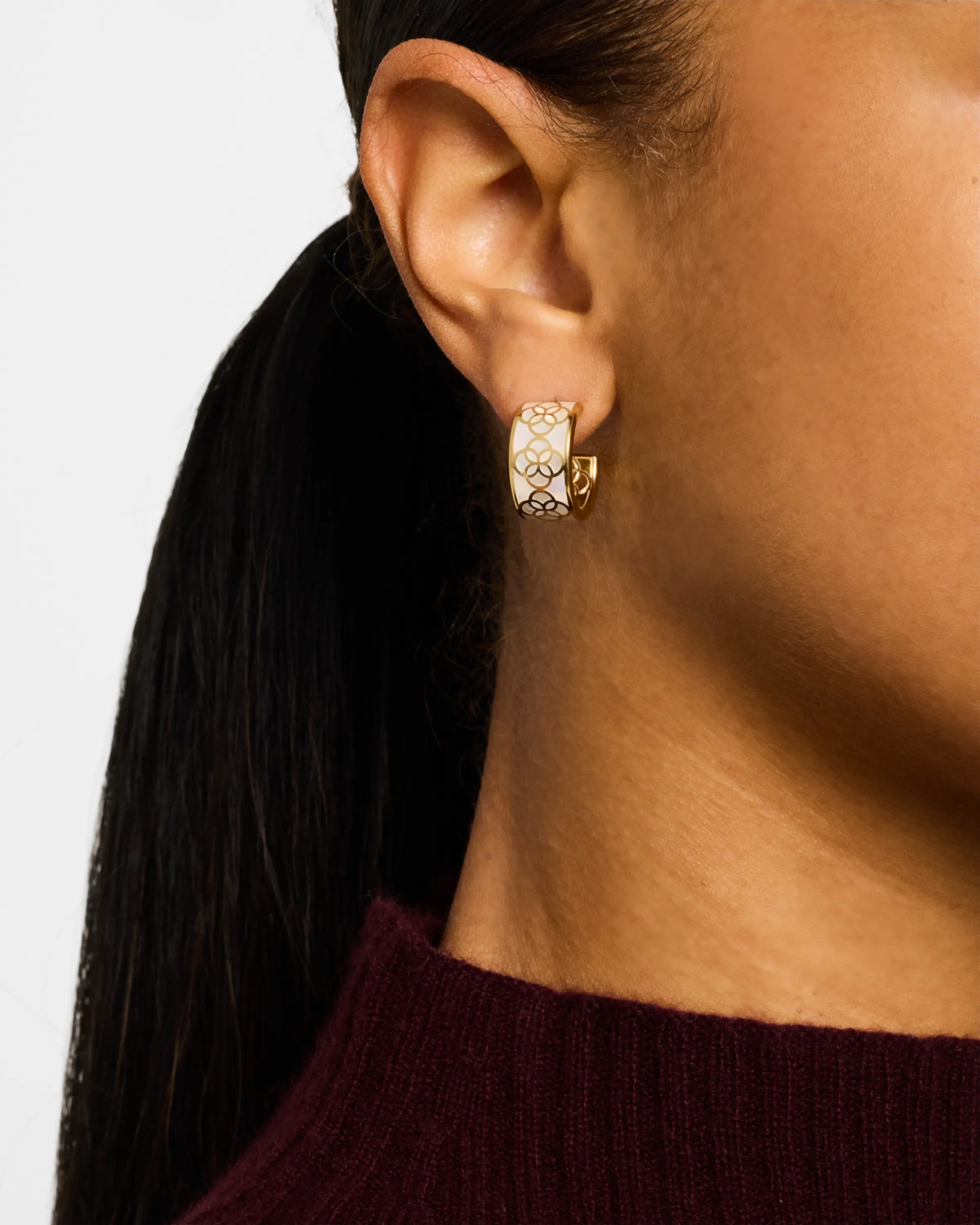 Close-up side profile of woman wearing gold vermeil sterling silver wide-band white mother of pearl floral lattice huggie hoop earring on ear with dark ponytail over burgundy top, Jan Leslie