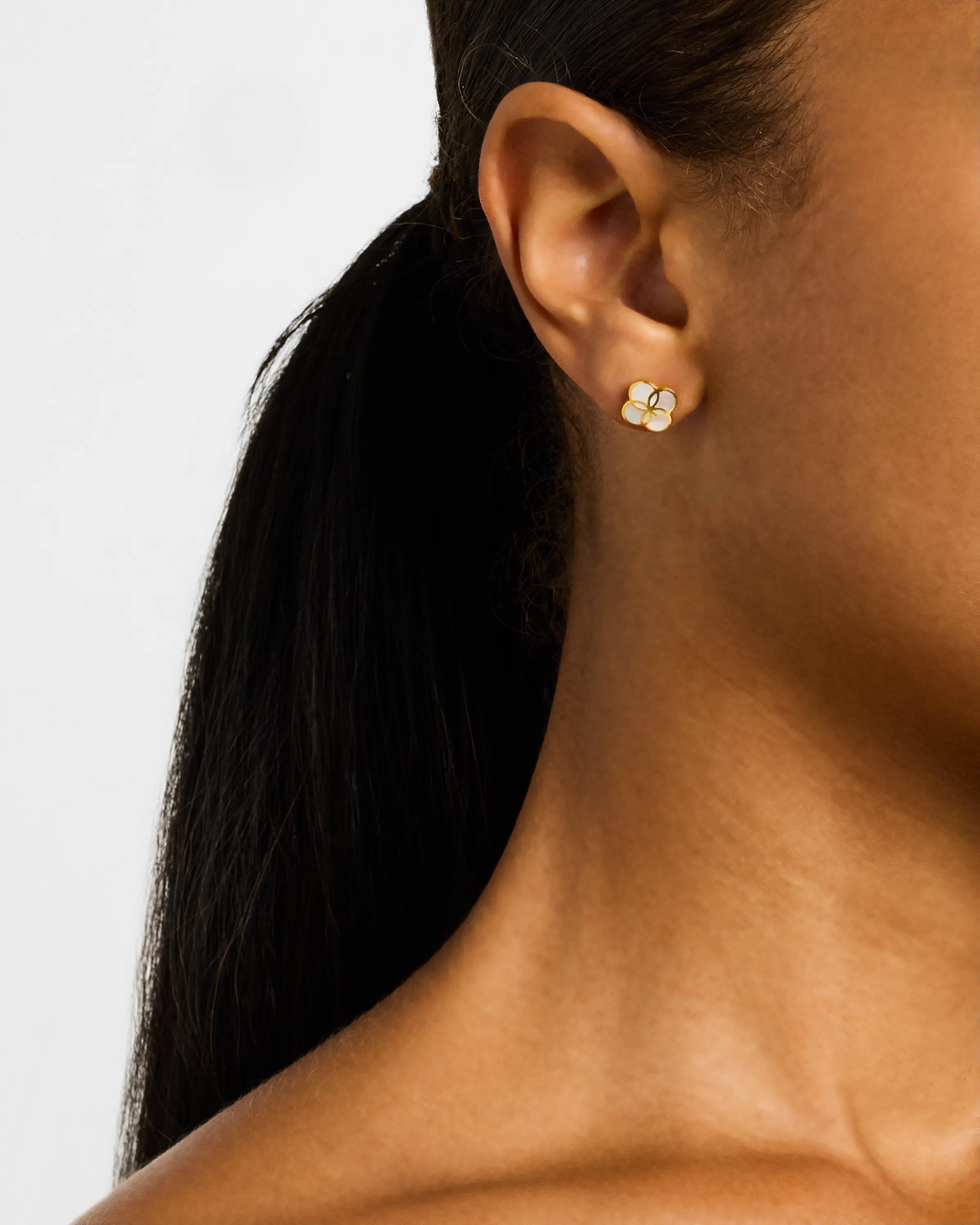 Woman wearing gold quatrefoil flower stud earrings with white mother of pearl inlay.