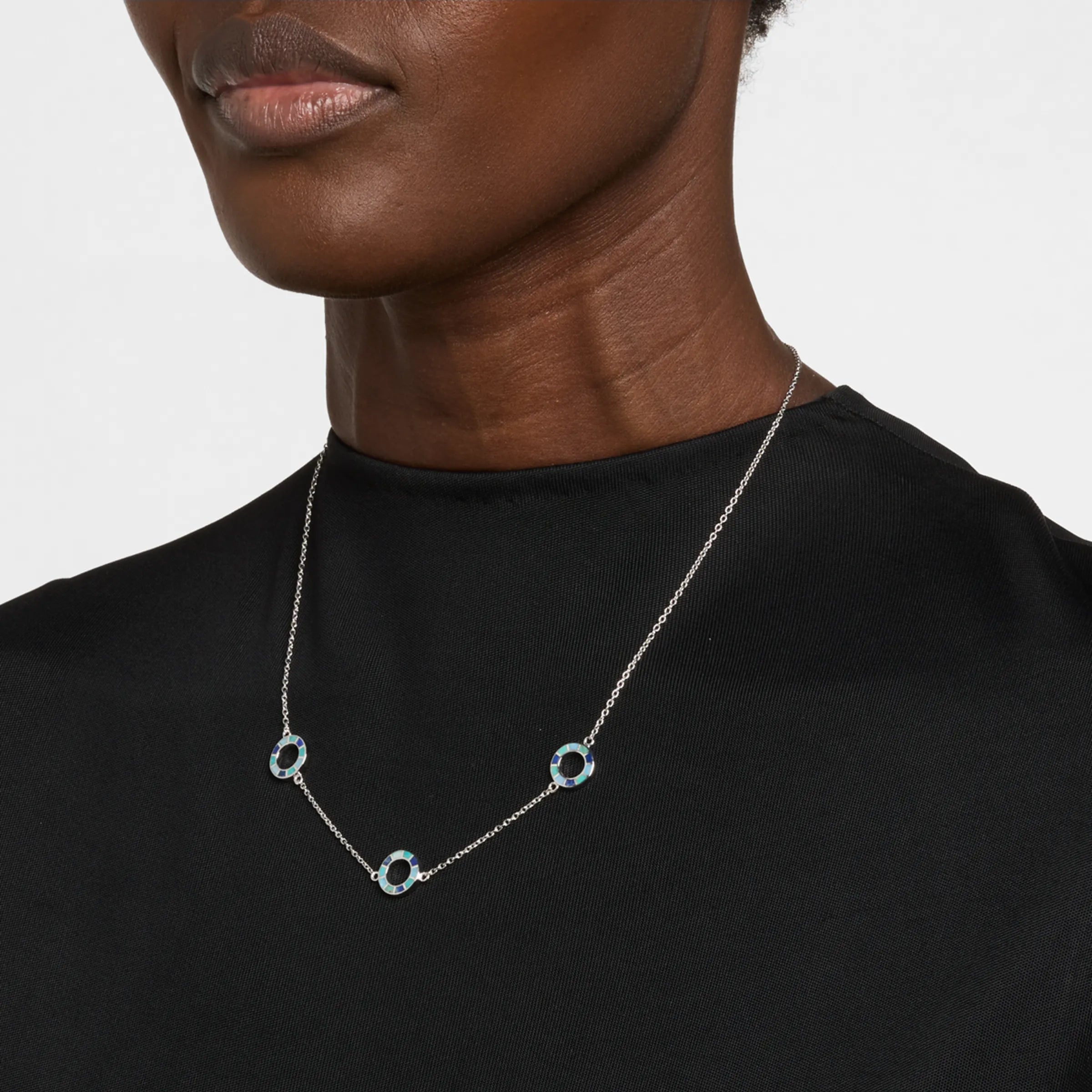 Woman in black crew neck top wearing a sterling silver three-station necklace with open circle blue and teal enamel mosaic pendants.