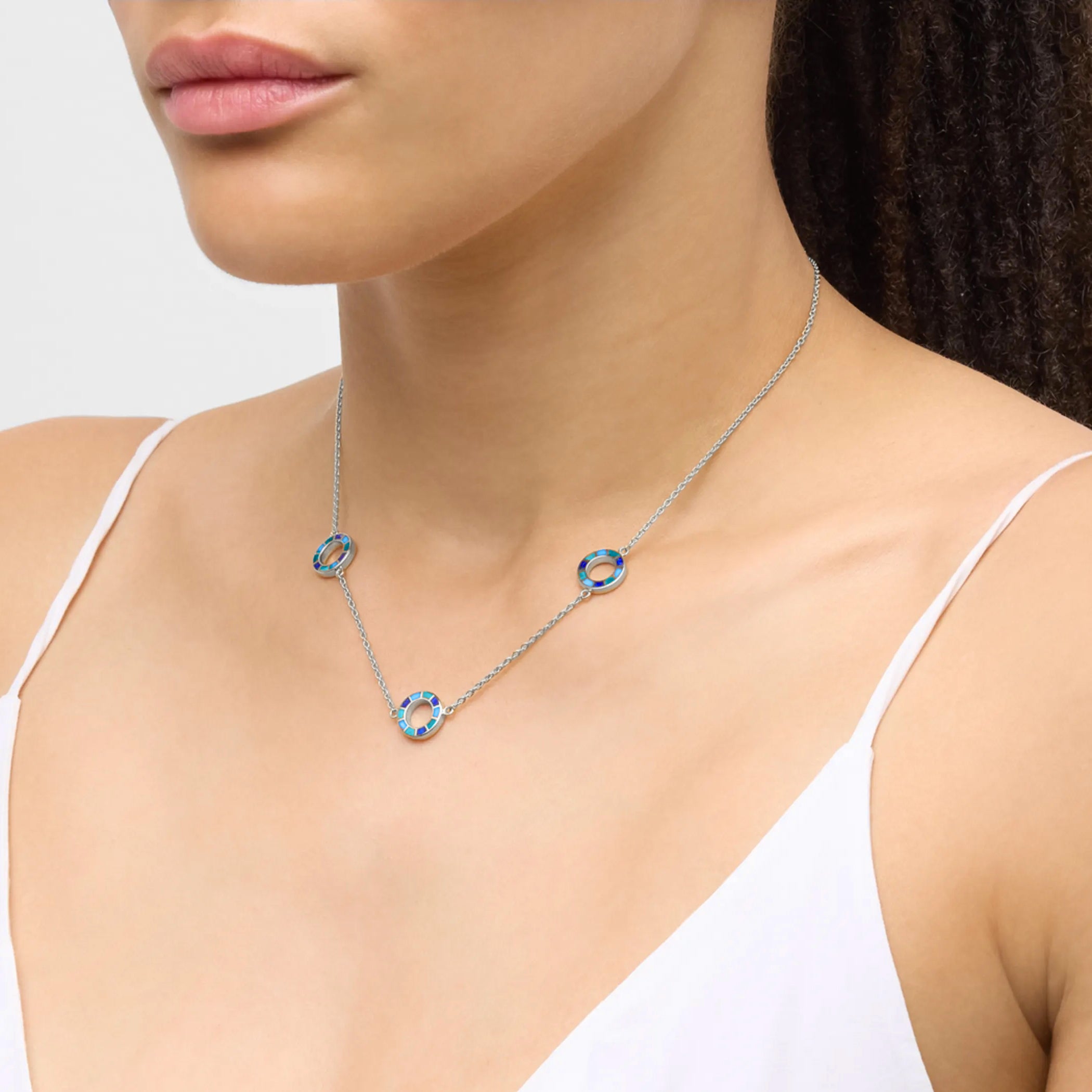 Woman in white camisole wearing a sterling silver three-station necklace with open circle blue and teal enamel mosaic pendants.