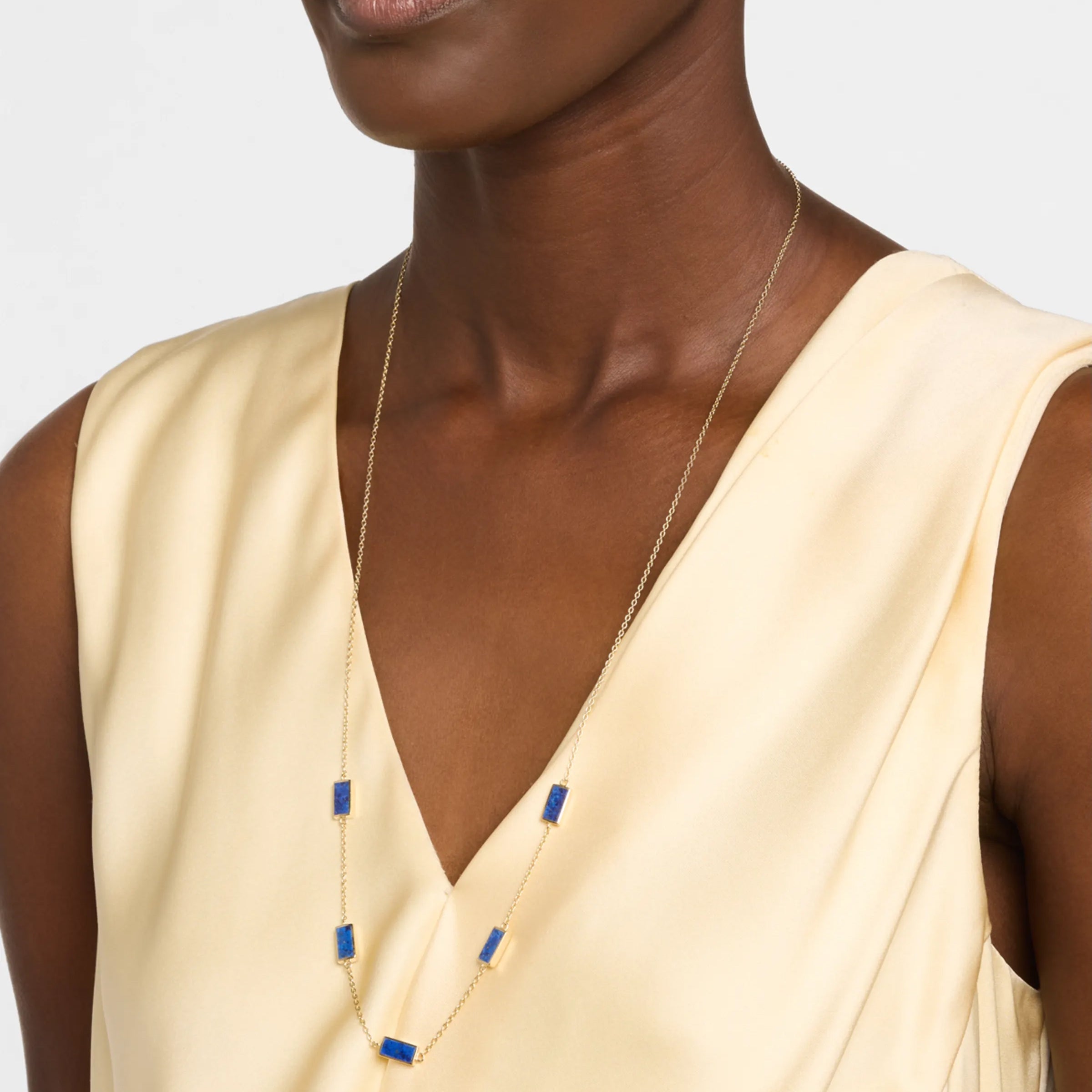 Woman in cream silk dress wearing a long gold chain station necklace with five rectangular lapis lazuli stone pendants.