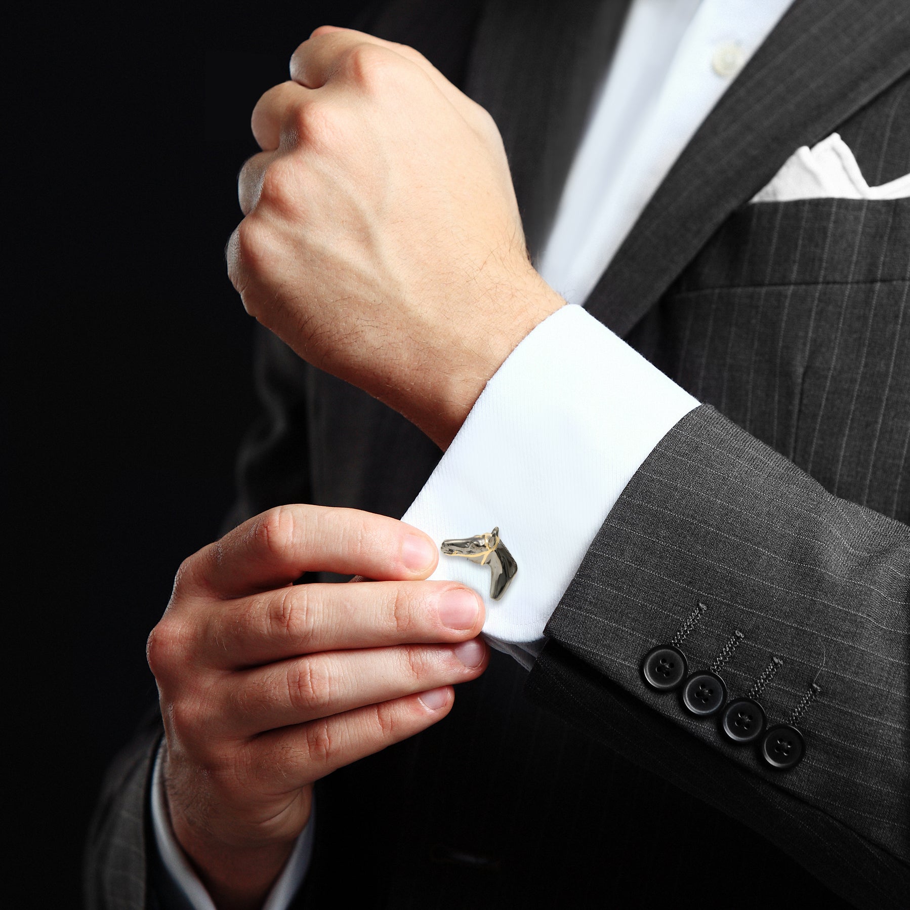 Man fastening gunmetal and gold 3D horse head cufflinks on white French cuff with charcoal pinstripe suit and white pocket square, luxury equestrian men's fine jewelry lifestyle.