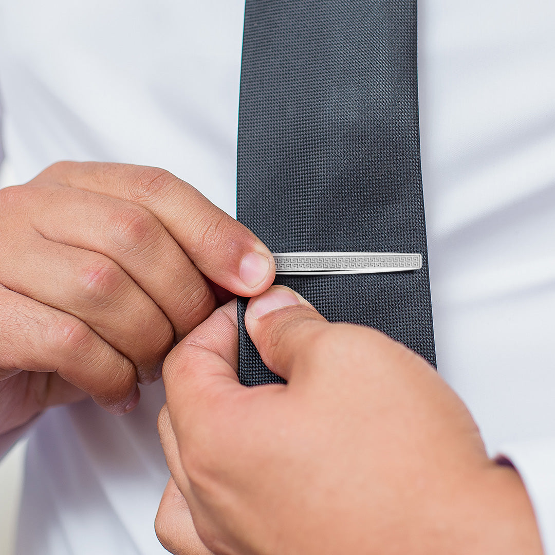 Photo of a male model placing the Greek Pattern Sterling Silver Tie Bar in matte on a dark grey tie. 